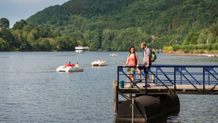 Ein Paar steht auf einem Steg am See in Einruhr, umgeben von Tretbooten und einem Ausflugsschiff. Im Hintergrund sind bewaldete Hügel zu sehen.