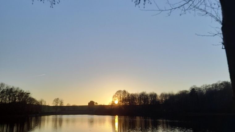 A calm lake under a clear sky at sunset. The reflection of light on the water creates a peaceful atmosphere.
