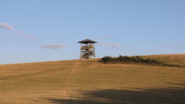 A lookout tower stands on a gentle hilltop. The sky is clear and blue, surrounded by a wide meadow.