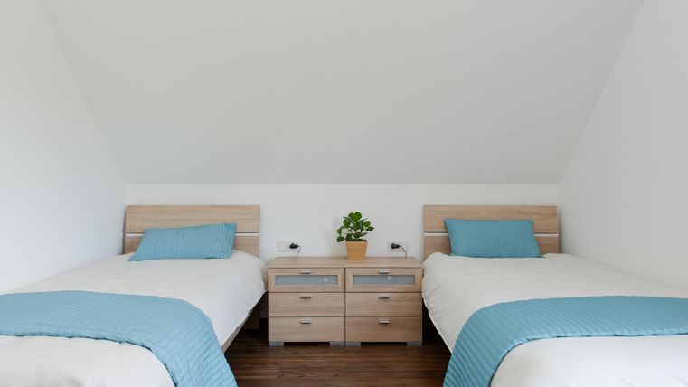 Ein gemütliches Schlafzimmer mit zwei Einzelbetten und hellen Holzmöbeln. Die Wände sind weiß und es gibt einen kleinen Pflanzenartikel auf dem Nachttisch.