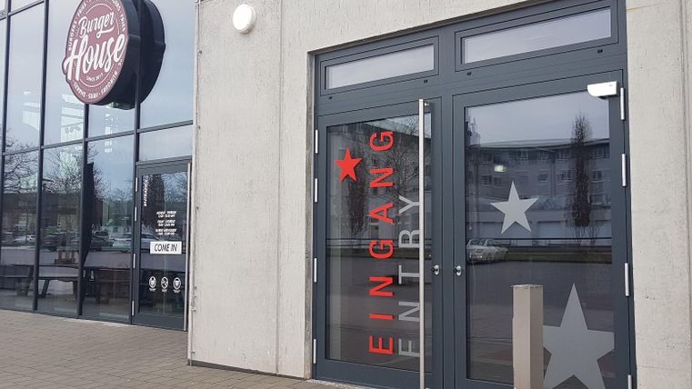 Eingangstür zu einem Restaurant mit dem Namen "Dinner House". Große Fenster zeigen die Außenansicht und eine moderne Fassade.