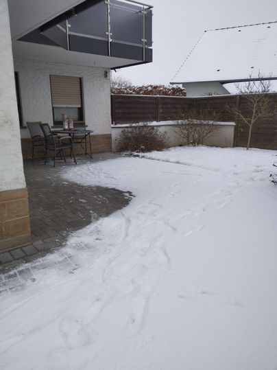 A snow-covered garden with footprints and a small seating area. In the background, houses and a cloudy sky are visible.