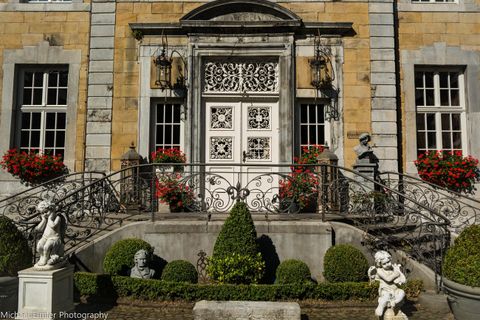 Eine prächtige Eingangstür mit kunstvollen Verzierungen und einer eleganten Treppe. Auf beiden Seiten sind dekorative Statuen und blühende Pflanzen zu sehen.