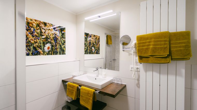 Ein modernes Badezimmer mit einem Waschbecken und Holzregal. Gelbe Handtücher und blühende Bilder verleihen dem Raum Farbe und Gemütlichkeit.