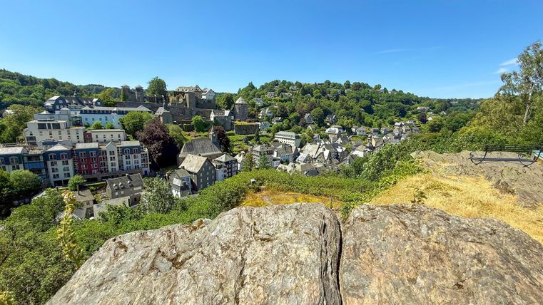 A picturesque view of a small village surrounded by green hills. In the foreground, you can see a large stone rock.
