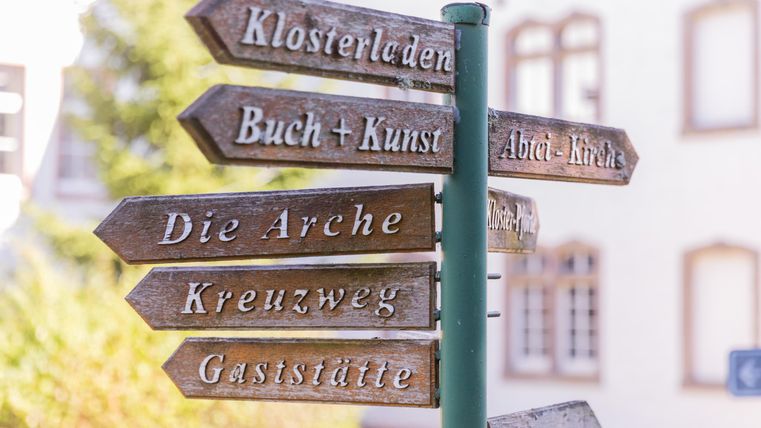 A signpost with various directions. The signs indicate places like "Monastery Shop" and "Books + Arts".