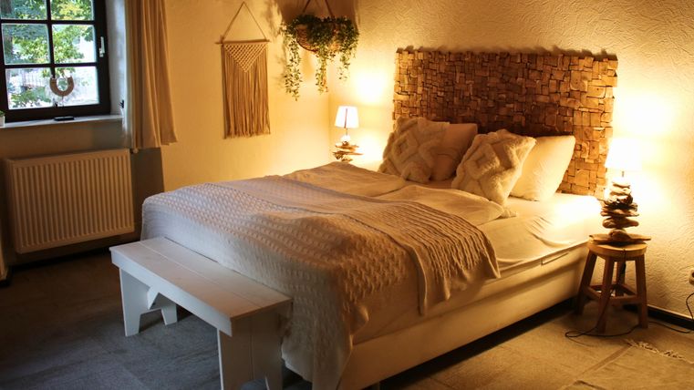 A cozy bedroom with a large bed and warm light. The walls are in soft colors and there are decorative plants.