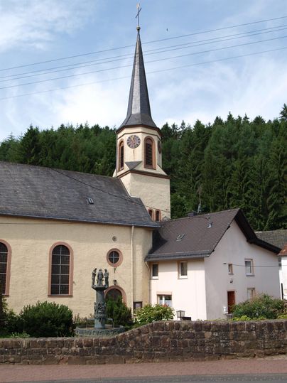 Eine alte Kirche mit einem hohen Turm steht neben einem kleinen Gebäude. Im Hintergrund sind Bäume zu sehen.
