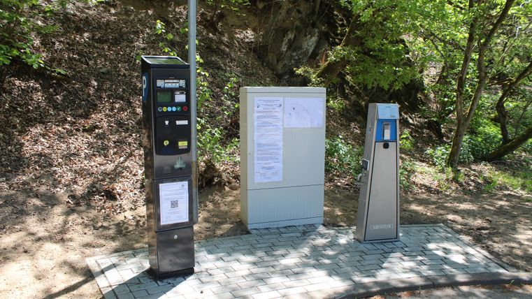Drei Parkautomaten stehen an einem Waldweg. Im Hintergrund sind grüne Bäume sichtbar.