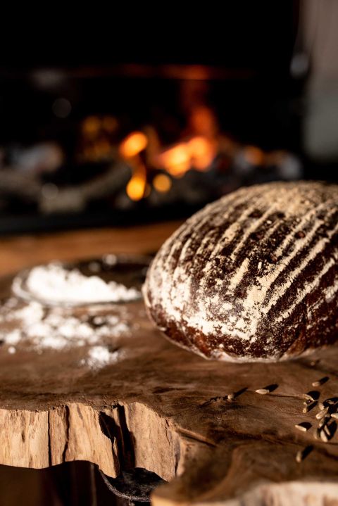 Een vers gebakken brood ligt op een houten plank. Op de achtergrond is een haardvuur te zien.