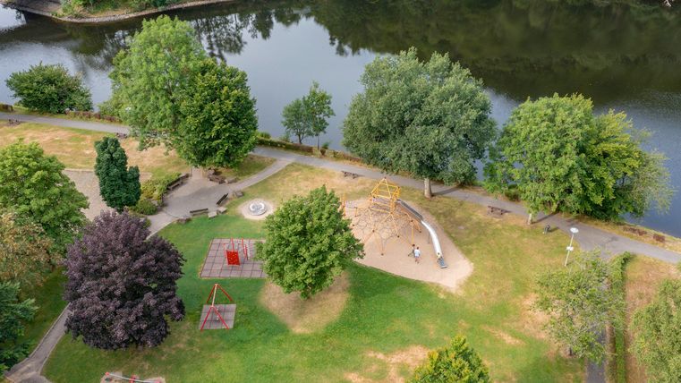 Een groene park aan het water met veel bomen. In het park zijn speeltoestellen en zitgelegenheden.