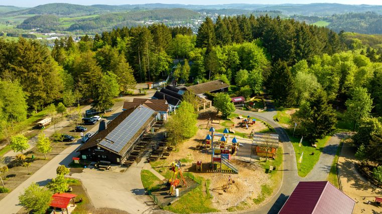 Een luchtige blik op een speelplaats omringd door bomen en hutten. De vriendelijke omgeving nodigt gezinnen uit tot ontspanning en spel.