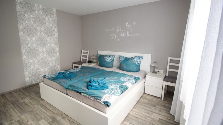 A modern bedroom with a large bed and cozy blue bedding. The walls are neutral, and there are two chairs next to the bed.