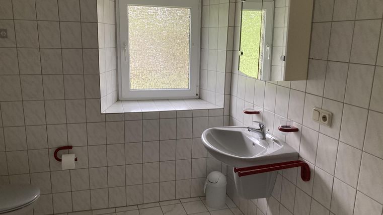 Een eenvoudige badkamer met witte tegels en rode accenten. Er is een raam, een wastafel en een toilet.
