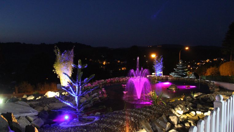 Ein schöner Garten mit beleuchteten Wasserfontänen und grünen Bäumen in der Dämmerung. Farbenfrohe Lichter spiegeln sich im Wasser und schaffen eine entspannende Atmosphäre.