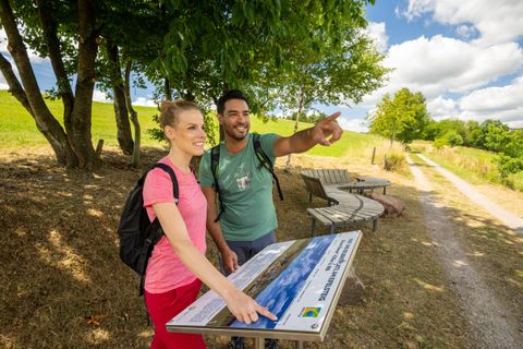 Zwei Wanderer stehen an einem Informationsschild im Freien. Sie schauen in die Ferne und scheinen einen schönen Tag in der Natur zu genießen.