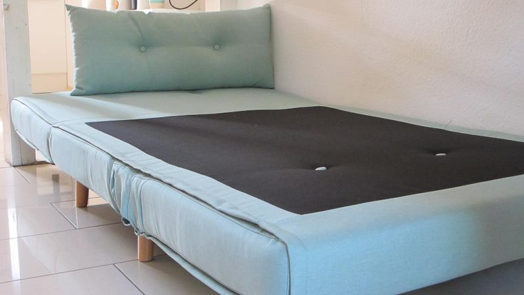 Ein modernes Bett mit einer blauen Matratze und einem schwarzen Mittelteil. Die Kopfstütze ist ebenfalls in hellblau und es steht auf vier Holzbeinen.