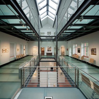 A modern interior with a glass roof and multiple levels. Various artworks hang on the walls, while seating arrangements are placed in the foreground.