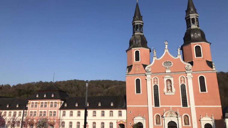 Eine beeindruckende Kirche mit zwei spitzen Türmen und einer rosa Fassade. Im Hintergrund sind historische Gebäude und grüne Hügel zu sehen.
