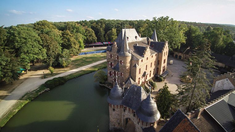 Een indrukwekkend kasteel met torens, omringd door een gracht. Het groene landschap en de bomen creëren een pittoreske sfeer.