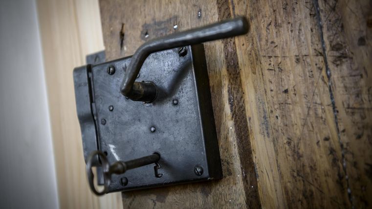 Ein alter, schwarzer Türverschluss aus Metall an einer Holzoberfläche. Der Verschluss hat einen einfachen Hebelmechanismus.