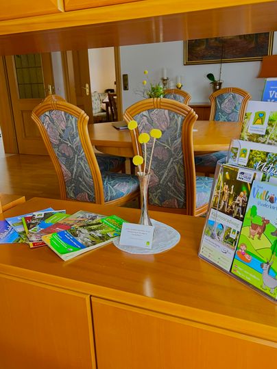 An inviting room with a wooden desk and a large dining table in the background. On the desk, there are informational brochures and a small vase with yellow flowers.