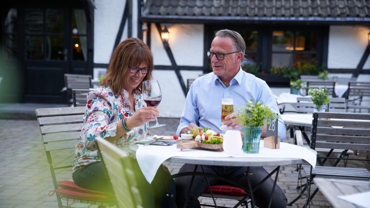 Ein Paar sitzt gemütlich im Freien und genießt ein Abendessen. Sie stoßen mit Wein und Bier an und wirken glücklich.