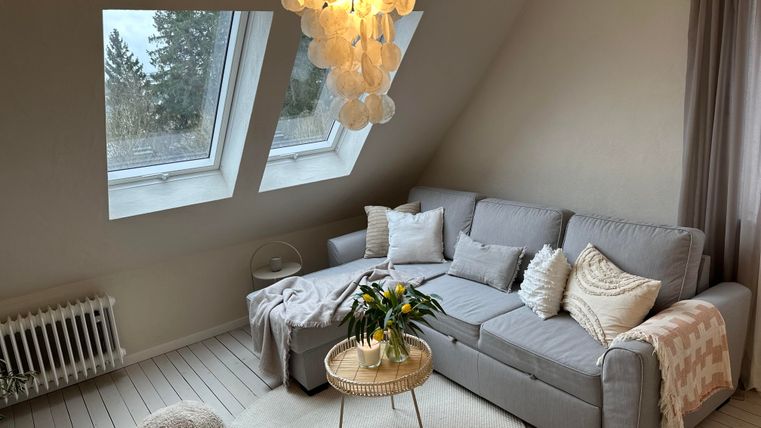 Een gezellige woonkamer met een grijze bank en een ronde salontafel. Grote ramen laten veel daglicht binnen en de decoratie is modern en uitnodigend.