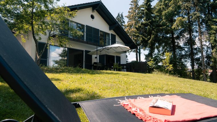 Ein modernes Haus umgeben von Bäumen. Im Vordergrund liegt eine Liege mit einem bunten Tuch und einem Buch.