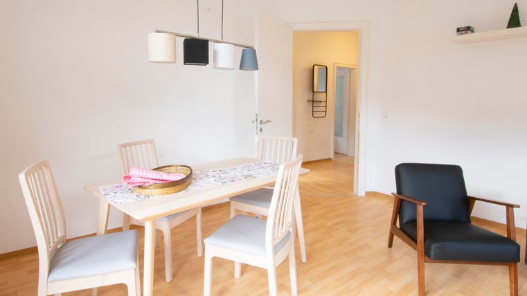 Ein modern eingerichteter Essbereich mit einem Tisch und vier Stühlen. In der Ecke steht ein schwarzer Sessel, und das Zimmer hat helle Wände und einen warmen Holzboden.