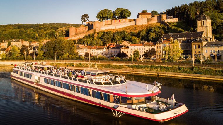 A ship is sailing on a river along a beautiful landscape. In the background, a castle and old buildings can be seen.