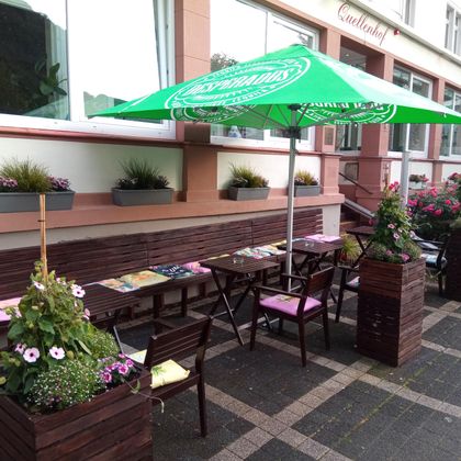 Ein einladender Außenbereich eines Cafés mit Holzmöbeln und einem grünen Sonnenschirm. Blumenkästen und Pflanzen schaffen eine angenehme Atmosphäre.