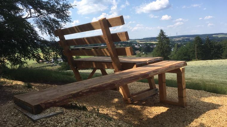 An oversized wooden bench stands on a hill overlooking the landscape near Kerschenbach under a blue sky with clouds.