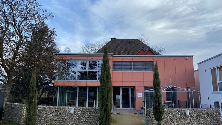 Ein modernes Gebäude mit einer pinken Fassade und großen Fenstern. Der Eingangsbereich ist von Bäumen und einem Steinzaun umgeben.