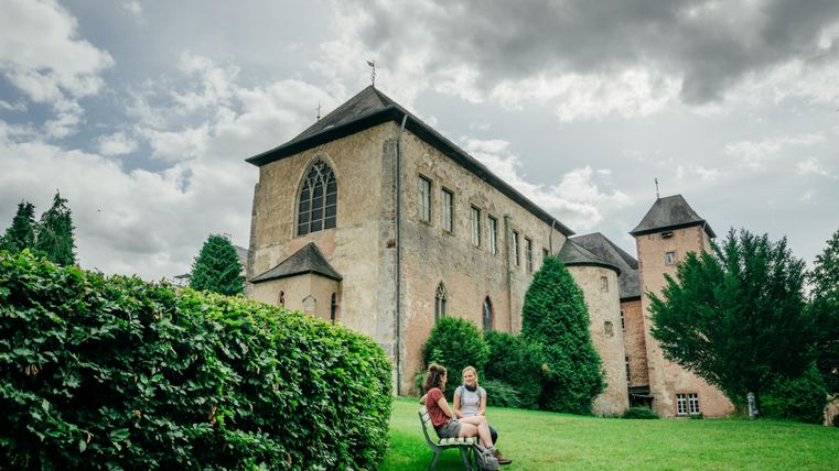 Eine alte Kirche umgeben von Grünflächen und Bäumen. Zwei Personen sitzen entspannt im Vordergrund auf einem Rasen.