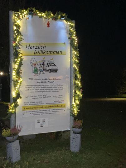 Ein beleuchtetes Willkommensschild, geschmückt mit Lichtern und Pflanzen. Es lädt zum Besuch eines Weihnachtsmarktes ein.
