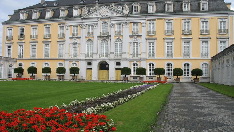 Ein beeindruckendes Schloss mit einer eleganten Fassade in Gelb und weißen Details. Im Vordergrund sind blühende Blumenarrangements zu sehen, umgeben von einem gepflegten Garten.
