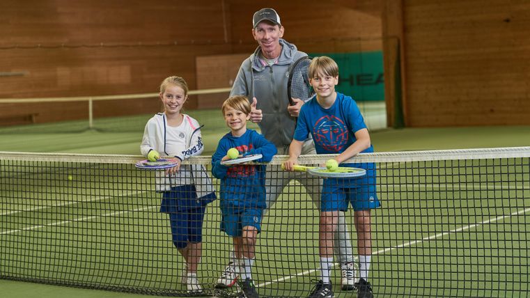 Eine Gruppe von drei Kindern steht an einem Tennisnetz, während ein Trainer hinter ihnen lächelt. Alle halten Tennisschläger und Bälle in der Hand.