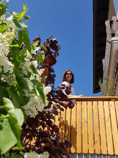 Een persoon staat op een balkon en kijkt glimlachend in de camera. Op de voorgrond zijn bloeiende planten te zien, en de lucht is helder en blauw.