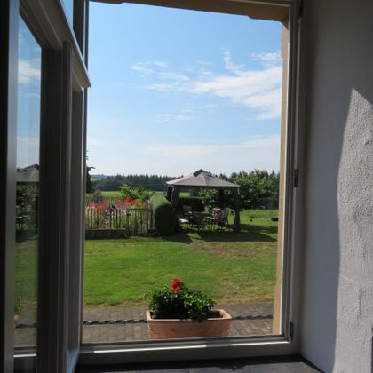 Ein Blick aus einem Fenster auf einen Garten mit einer Wiese und einem Pavillon. Der Himmel ist klar und blau, umgeben von Bäumen und bunten Blumen.