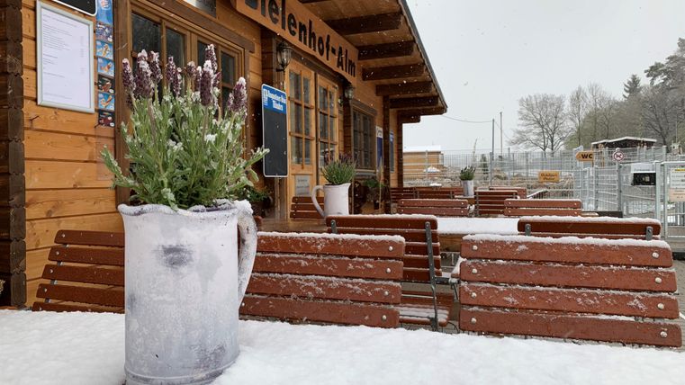 Sneeuwbedekte terras van de Bielenhof Alm met houten banken, een plantenbak en een houten gebouw op de achtergrond.