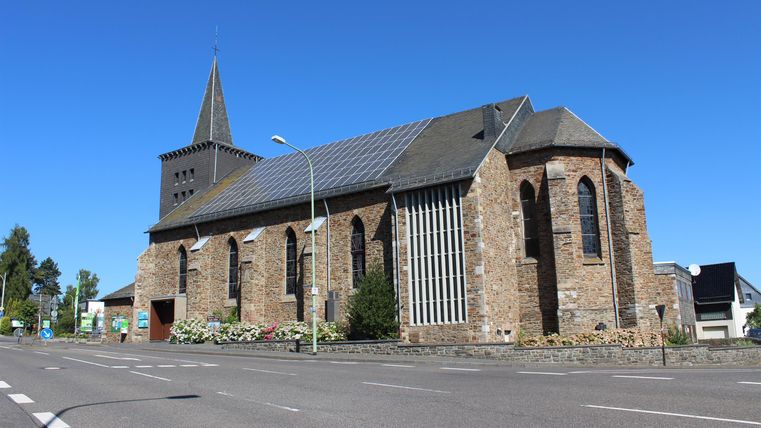 An old stone church with a pointed tower stands by a road. The sky is clear and blue.