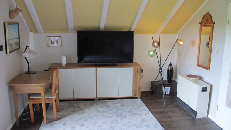 Ein gemütliches Wohnzimmer mit einem großen Fernseher und einem Schreibtisch. Die Wände sind hell und im Raum stehen farbenfrohe Leuchten.