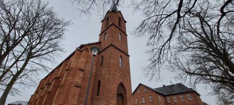 Eine rote Backsteinkirche mit einem hohen Turm steht im Vordergrund. Umgeben von Bäumen unter einem grauen Himmel.