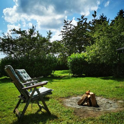 Een gezellige tuin met een stoel en een klein kampvuur. Omringd door bomen en groen gras onder een blauwe lucht.