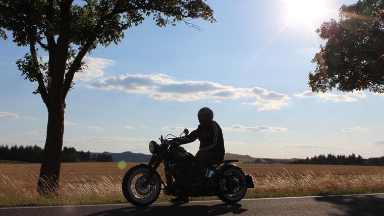 A motorcyclist is riding on a country road, surrounded by trees and a wide field. The sun is shining brightly in the sky, creating a relaxed atmosphere.