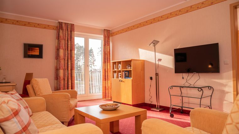 Een heldere woonkamer met comfortabele banken en een salontafel. Aan de muur hangt een televisie, en grote deurpuien zorgen voor veel natuurlijk licht.