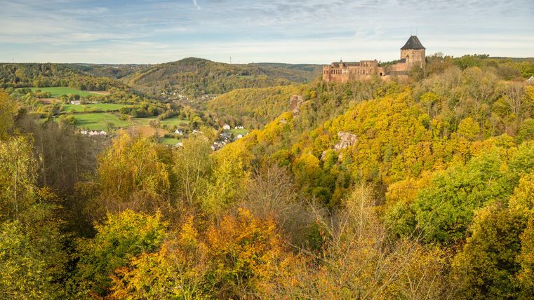 Eine beeindruckende Burgruine auf einem Hügel, umgeben von bunten Herbstbäumen. Im Hintergrund erstrecken sich sanfte Hügel und eine grüne Landschaft.