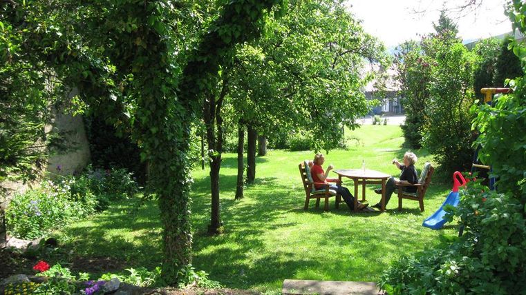 Ein sonniger Garten mit grünen Bäumen und einer gemütlichen Sitzecke. Zwei Personen entspannen sich am Tisch und genießen die Natur.