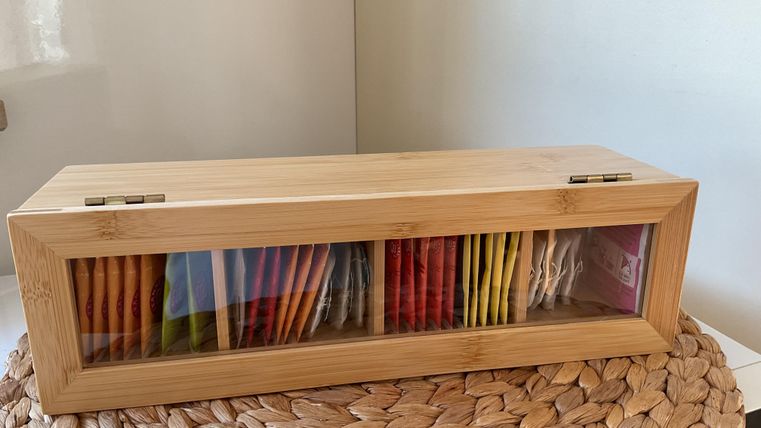 An elegant wooden box with a transparent window that contains various tea bags in bright colors. The box is placed on a woven background.